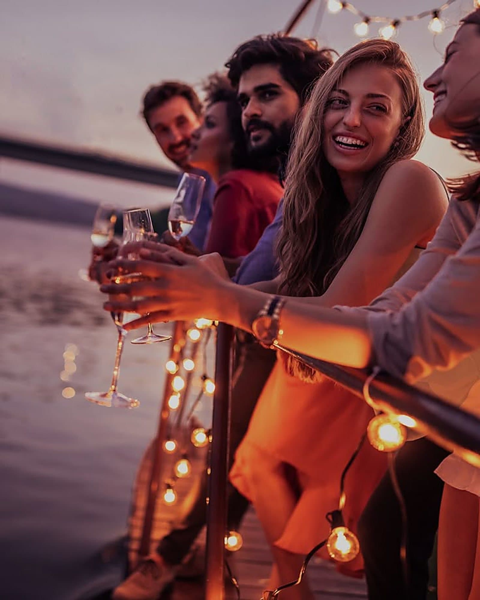Guests raising glasses on deck at sunset, with string lights along the rail